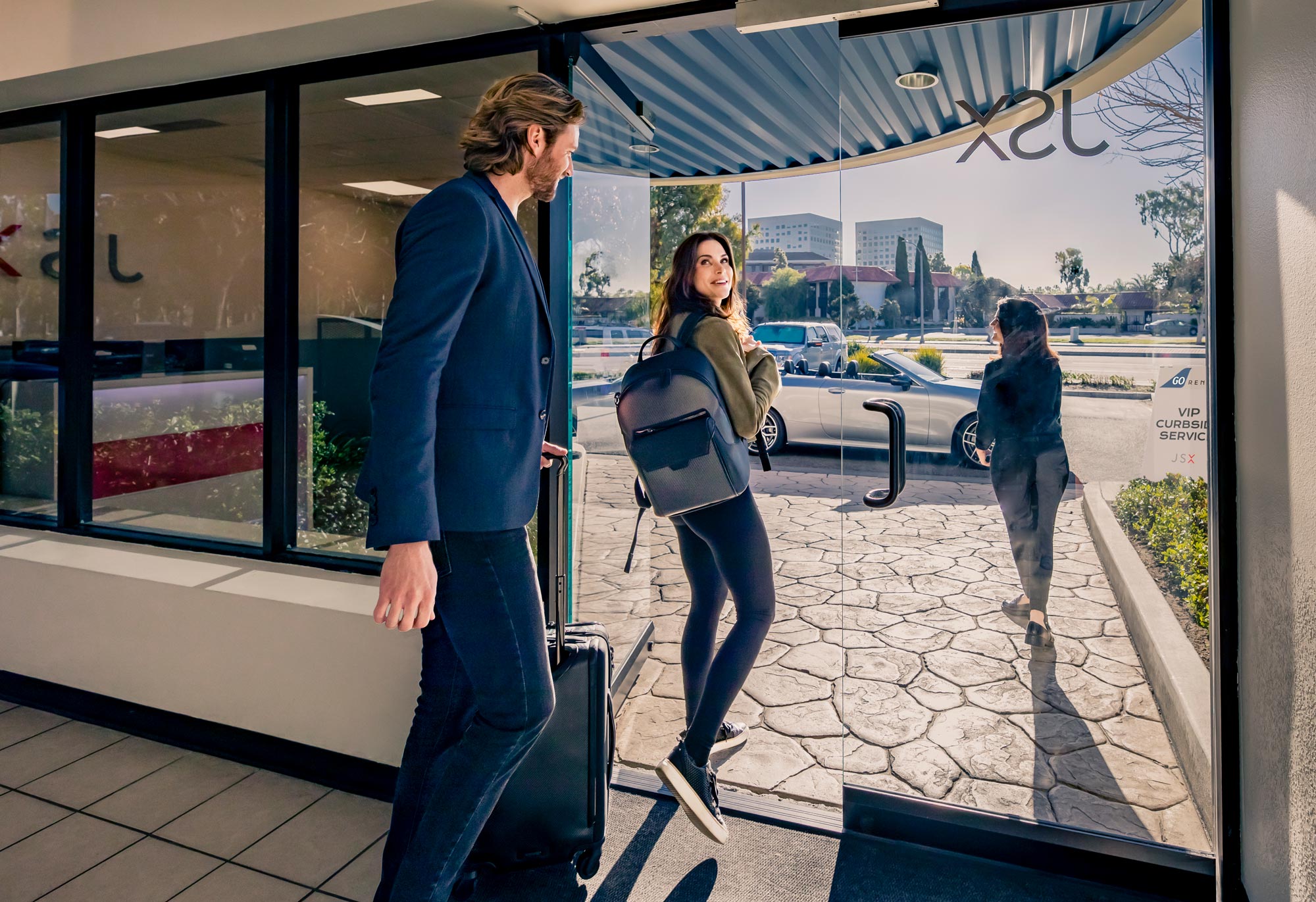 A man is holding his luggage suitcase while smiling at a woman going out of the door with her backpack.
